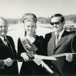 Four people (all of whom are white) posing and smiling for a ceremonial ribbon-cutting ceremony to mark the official opening of the Simi Freeway/SR 118 in 1970. A woman wearing a "Miss Simi Valley" sash stands in the middle of the group holding a large pair of scissors. The group stands in the middle of the freeway with a few cars and people in the background.